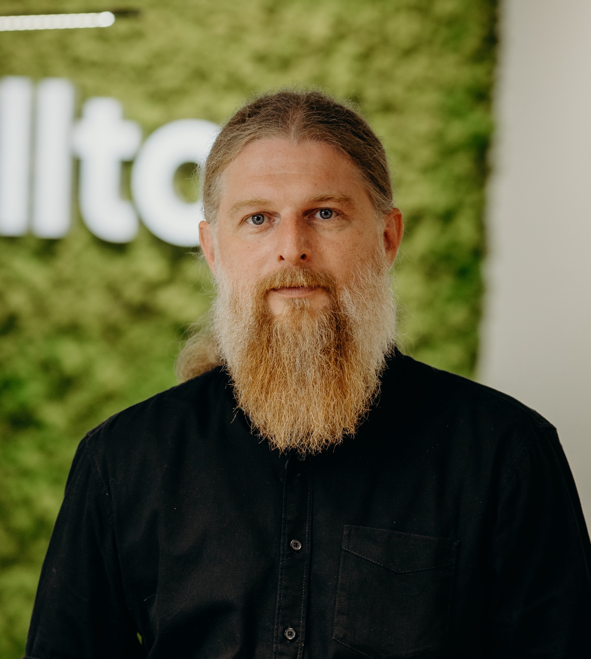 A man with a long beard and long hair wearing a black shirt, standing in front of a green moss wall with 'Kellton' logo visible.